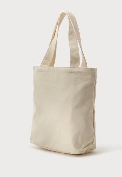 Sac fourre-tout en toile beige uni avec deux larges bretelles et une base rectangulaire, présenté sur fond blanc.