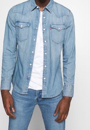 Hombre con camisa de mezclilla azul claro con botones de presión, sobre una camiseta blanca, combinada con jeans a juego del mismo tono azul claro.