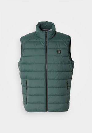 Colete verde acolchoado com gola alta, fecho frontal e dois bolsos laterais; apresenta costura horizontal e um pequeno logótipo no peito.