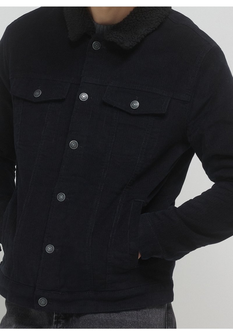 Veste en velours côtelé bleu marine avec un col en fourrure noire douce. Comprend deux poches poitrine, une fermeture à boutons et un tissu texturé pour la chaleur.