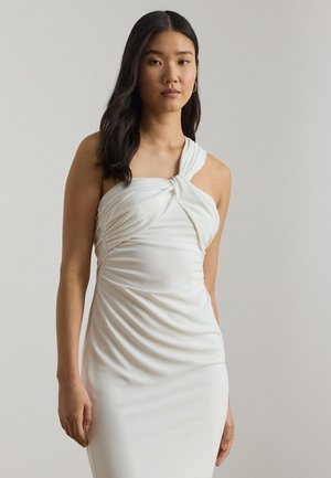 Noor naine, kellel on pikad tumedad juuksed, seljas valge ühe õlaga kroogitud kleit, seisab lihtsa hele tausta ees.