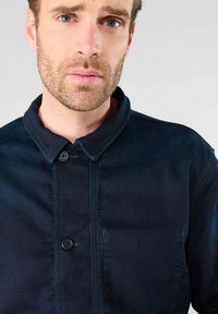 Marineblauwe denimshirt met kraag, knopen aan de voorkant en een borstzak. Heeft zichtbaar stiksel en een gladde textuur.