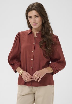 Junge Frau mit langen braunen Haaren, die eine rostfarbene Bluse mit Knöpfen und aufgepufften Ärmeln sowie beige Hosen trägt, ergänzt durch goldenen Schmuck.