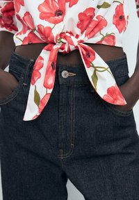 Blusa floreale bianca con papaveri rossi, legata in vita. Jeans in denim scuro con cuciture gialle e due tasche frontali.