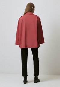 Giacca oversize rossa scura con colletto e spalline, abbinata a pantaloni neri slim-fit e stivaletti alla caviglia. Tessuto dalla superficie liscia.