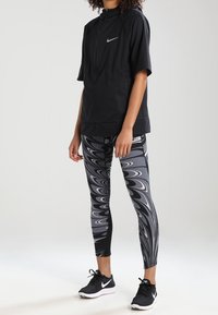 Černý vrchní díl s krátkými rukávy a polovičním zipem, doplněný o upnuté legíny s vířivým černobílým vzorem a černými sportovními botami.