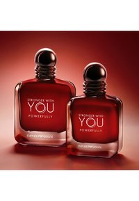 Deux flacons de parfum en verre teinté rouge avec des bouchons ronds, étiquetés "Stronger With You Powerfully" par Emporio Armani sur un fond dégradé rouge assorti.