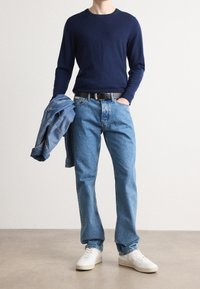 Maglione blu navy, jeans di denim blu chiaro, cintura nera, tiene una giacca di denim e sneakers bianche. Outfit casual con vestibilità rilassata e design classico.