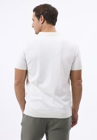 Kurzärmliges, cremefarbenes Poloshirt aus weichem Stoff. Taillierte Passform mit geripptem Kragen und Saum. Sieht auf einem Model von hinten aus.
