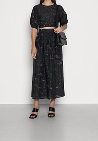 Abito maxi con stampa floreale in tessuto scuro, con maniche a sbuffo e vita stretta. Abbinato a sandali neri open-toe e a una borsa a spalla nera.