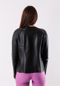 Giacca di pelle nera con un design aderente, colletto rotondo e dettagli delle cuciture. Modello indossato con leggings viola, visto da dietro.