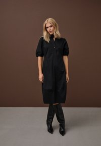 B blondinė moteris juoda kelio ilgio suknele su pūstomis rankovėmis ir juodais, aukštais, smailiais bateliais, stovinti prie rudos spalvos fono.