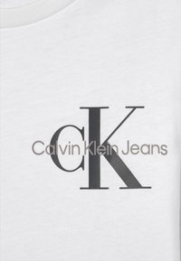 Biała bawełniana koszulka z dużą czarną literą "K" z okrągłym akcentem, towarzyszącym napisem "Calvin Klein Jeans" w mniejszej szarej czcionce.