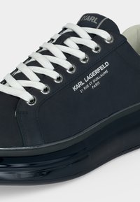 Közeli kép egy fekete Karl Lagerfeld sportcipőről fehér fűzőkkel, fényes fekete talppal és márkajelzéssel az oldalán és a nyelvén.