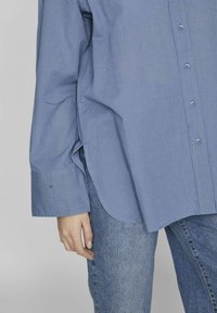 Svetlo modra srajca s dolgimi rokavi, s krivuljo na dnu in subtilno teksturo. Ujemajoče se oprijete modre jeans hlače.