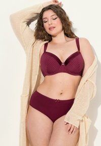 Burgundin rintaliivit, joissa on pilkkuja ja pehmeät kupit, yhdistettynä vastaaviin pikkupöksyihin, joissa on nauha koristeena. Yllä vaaleanbeige neulecardigan.