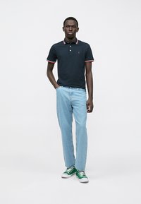 Ναυτικό πόλο μπλουζάκι με κόκκινες και άσπρες λεπτομέρειες, ανοιχτό μπλε τζην και πράσινα αθλητικά. Το μοντέλο στέκεται μπροστά σε λευκό φόντο.