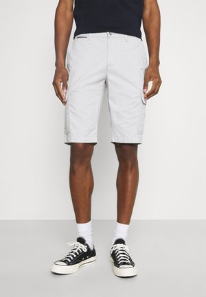 Man in lichtgrijze cargoshorts, witte sokken, zwarte low-top sneakers en een zwart shirt, staande tegen een effen achtergrond.