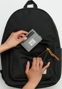 Svart tygryggsäck med en framficka. Hand placerar en grå plånbok med texturerad yta och en logotyp på fickans dragkedjesida.