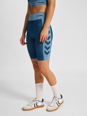 Vrouw draagt blauwe hummel sportshorts en sportbeha met witte sokken en witte sneakers, staand tegen een effen achtergrond.