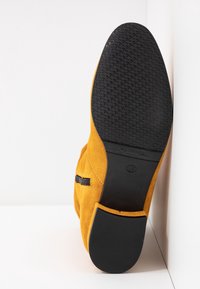 Bottine en suede jaune avec une semelle en caoutchouc noir texturé, dotée d'une fermeture éclair sur le côté et d'une finition lisse.