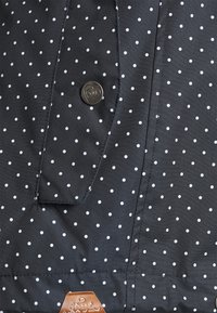 Svart tyg med vita prickar har en metallknapp och en brun läderetikett med präglad text. Slät textur och figurnära design.