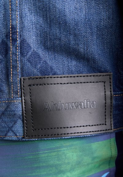 Traper jakna od tamnoplave tkanine, s narančastim šavovima, i pravokutnom crnom kožnom oznakom s utisnutim natpisom "Ahluwalia." Ima teksturiranu površinu.