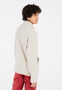 Chaqueta de forro polar en beige claro, con un cuello alto, superficie texturizada y bolsillos laterales, combinada con pantalones rojos para un contraste brillante.