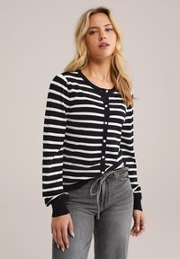 Cardigan rayé noir et blanc en tissu tricoté, avec des boutons en perles et des poignets côtelés, associé à un pantalon gris taille haute.