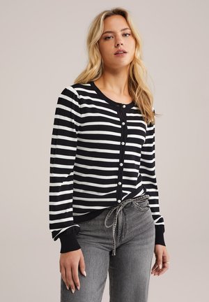 Cardigan rayé noir et blanc en tissu tricoté, avec des boutons en perles et des poignets côtelés, associé à un pantalon gris taille haute.