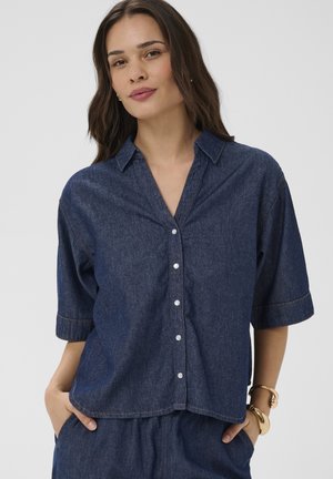 Kobieta ubrana w luźną ciemną koszulę z denimu z krótkimi rękawami na guziki oraz pasujące spodnie, z rękami w kieszeniach, zwrócona przodem.