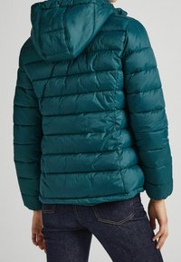 Teal pufferjacka gjord av glänsande material med horisontell quiltad mönster och huva, i kombination med mörkblå jeans.