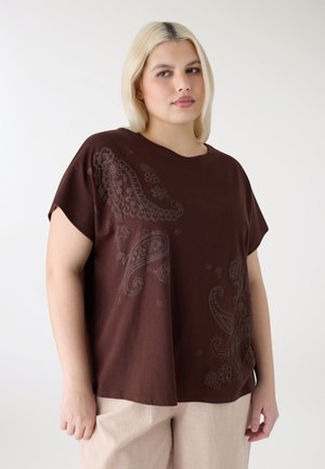 Giovane donna con capelli biondi che indossa una maglietta marrone larga con motivi paisley e pantaloni beige chiaro, in piedi contro uno sfondo semplice.