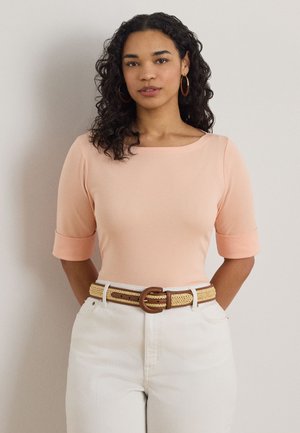 Tânără cu păr creț și închis la culoare, purtând un top piersică cu mâneci până la cot, pantaloni albi, o curea țesută și cercei mari rotunzi, stând în fața unui perete deschis la culoare.