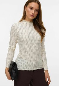 Crèmekleurige top met lange mouwen, voorzien van een gegolfde halslijn, gestructureerde stof en een subtiel patroon van meerkleurige stippen, gecombineerd met een zwarte clutch.