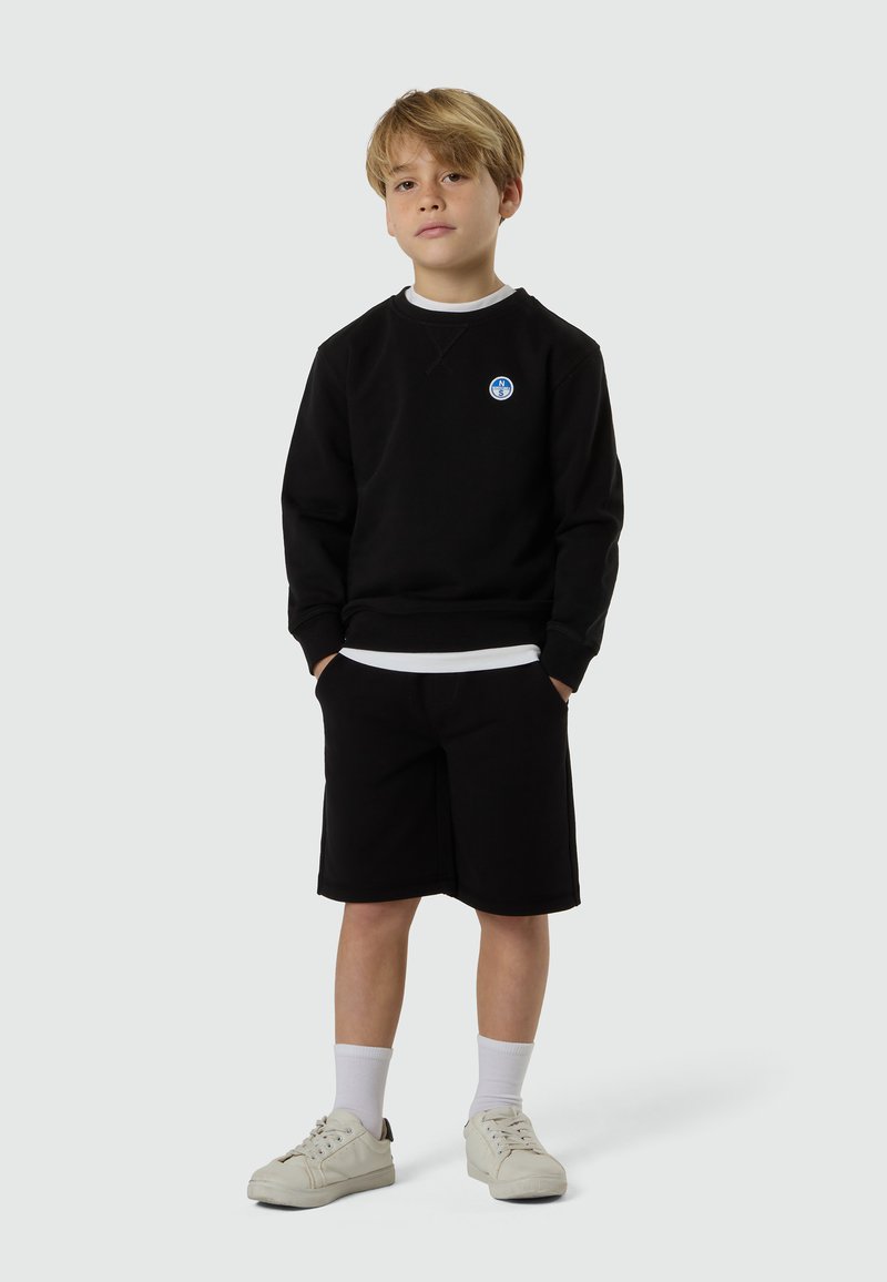 Ragazzo giovane con le mani nelle tasche, indossa una felpa nera con logo, pantaloncini neri, calzini bianchi e scarpe da ginnastica bianche, in piedi su uno sfondo semplice.