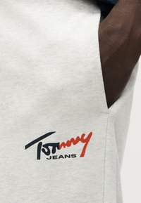 Κοντινό ενός χεριού στην τσέπη παντελονιού ανοιχτού γκρι με μαύρο και κόκκινο λογότυπο Tommy Jeans τυπωμένο κοντά στην τσέπη.