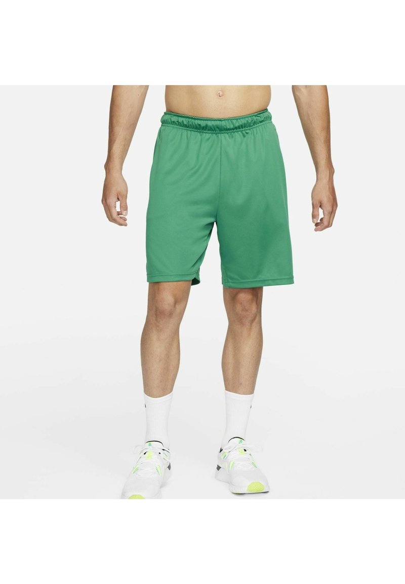 Homme portant un short de sport vert, des chaussettes blanches hautes et des baskets blanches, debout, les bras détendus le long du corps, sur un fond blanc.