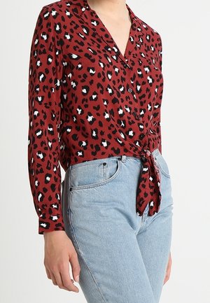 Vrouw draagt een rood blouse met luipaardprint en lange mouwen, gestrikt in de taille, gecombineerd met lichtblauwe, hooggesneden jeans.