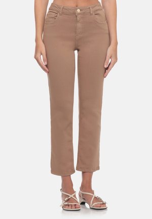 Donna che indossa pantaloni beige a gamba dritta e taglio corto e sandali bianchi con cinturini, in piedi davanti a uno sfondo semplice.
