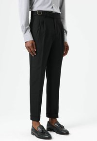 Zwarte, op maat gemaakte broek met plooien, voorzien van een riem, omslagzomen en een gladde textuur. Gecombineerd met glanzende zwarte loafers en een grijs overhemd.