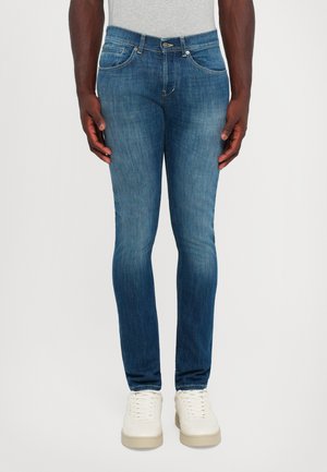 Uomo in piedi che indossa jeans slim-fit blu medio e sneaker bianche, con le mani rilassate ai lati, su uno sfondo chiaro e semplice.