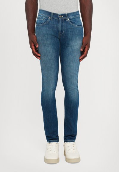 Uomo in piedi che indossa jeans slim-fit blu medio e sneaker bianche, con le mani rilassate ai lati, su uno sfondo chiaro e semplice.