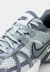 Sneaker grigio chiaro con materiale in rete, accenti in gomma e un prominente logo Nike Swoosh nero. Presenta allacciature tonali e sovrapposizioni testurizzate.