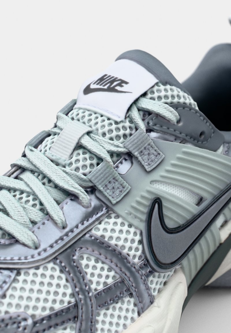 Zapatilla gris claro con material de malla, acentos de goma y un prominente Swoosh negro de Nike. Presenta un cordón tonal y superposiciones texturizadas.