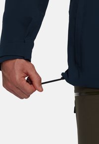 Veste imperméable marine avec cordon élastique au poignet, tissu texturé, main tenant le cordon. Pantalon vert partiellement visible.