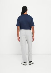 Osoba s dugom pletenicom stoji okrenuta leđima, odjevena u tamnoplavu majicu, svijetlosive trenirke i bijele tenisice s crnim detaljima.