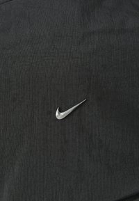 Musta kangaspinta, jossa on selkeä hopeinen Nike-swoosh-logo. Materiaali näyttää teksturoidulta ja siinä on hieman kiiltoa.