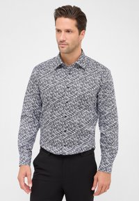 Uomo che indossa una camicia a maniche lunghe con bottoni, caratterizzata da un motivo geometrico bianco e nero, abbinata a pantaloni neri, in piedi davanti a uno sfondo neutro.