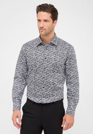 Mann, der ein langärmliges, knöpfbares Hemd mit schwarz-weißer geometrischer Musterung trägt, kombiniert mit schwarzen Hosen, steht vor einem schlichten Hintergrund.